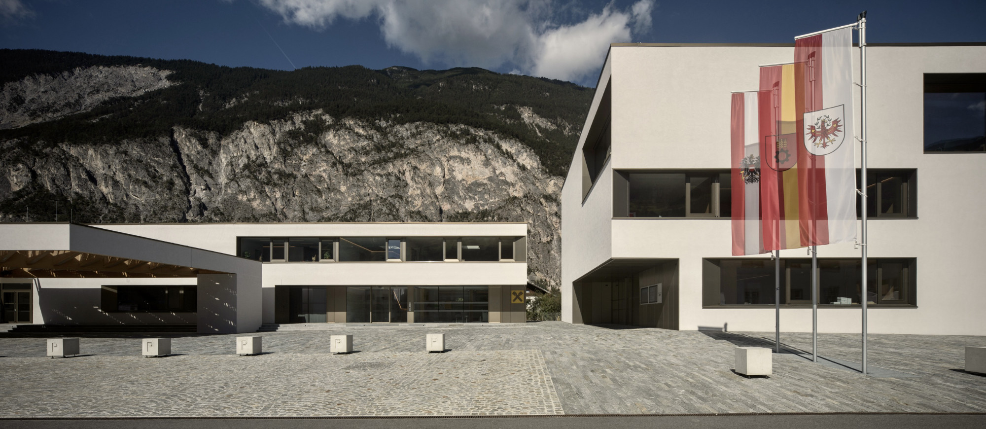 Gemeindezentrum Haiming, Gebäudeansicht mit Putzfassade, Fahnenmasten und Bergpanorama