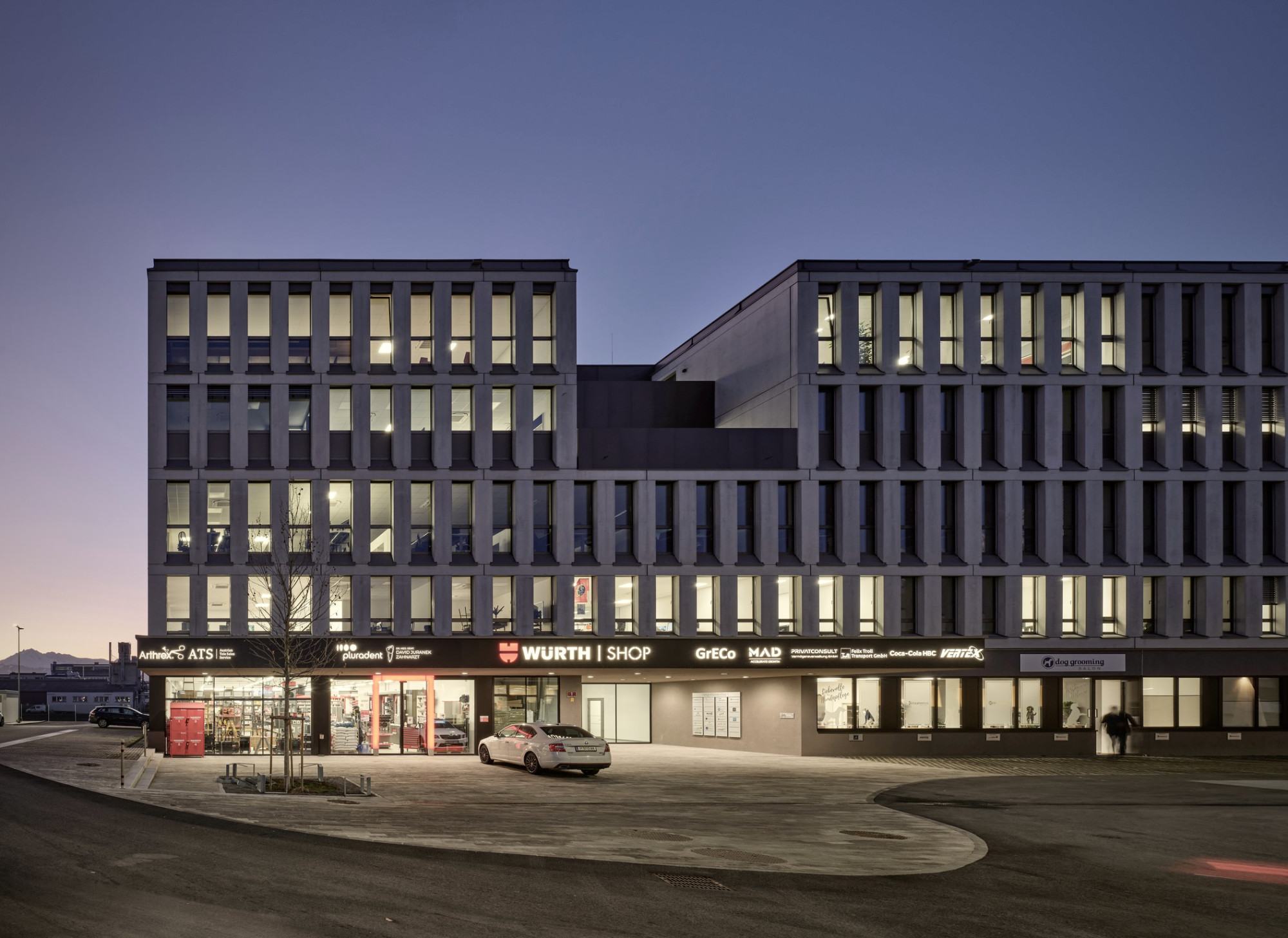 Helga Krismer Platz, Innsbruck, Gebäudeansicht bei Dämmerung mit beleuchteten Fensterreihen und Geschäftszeile im Erdgeschoss